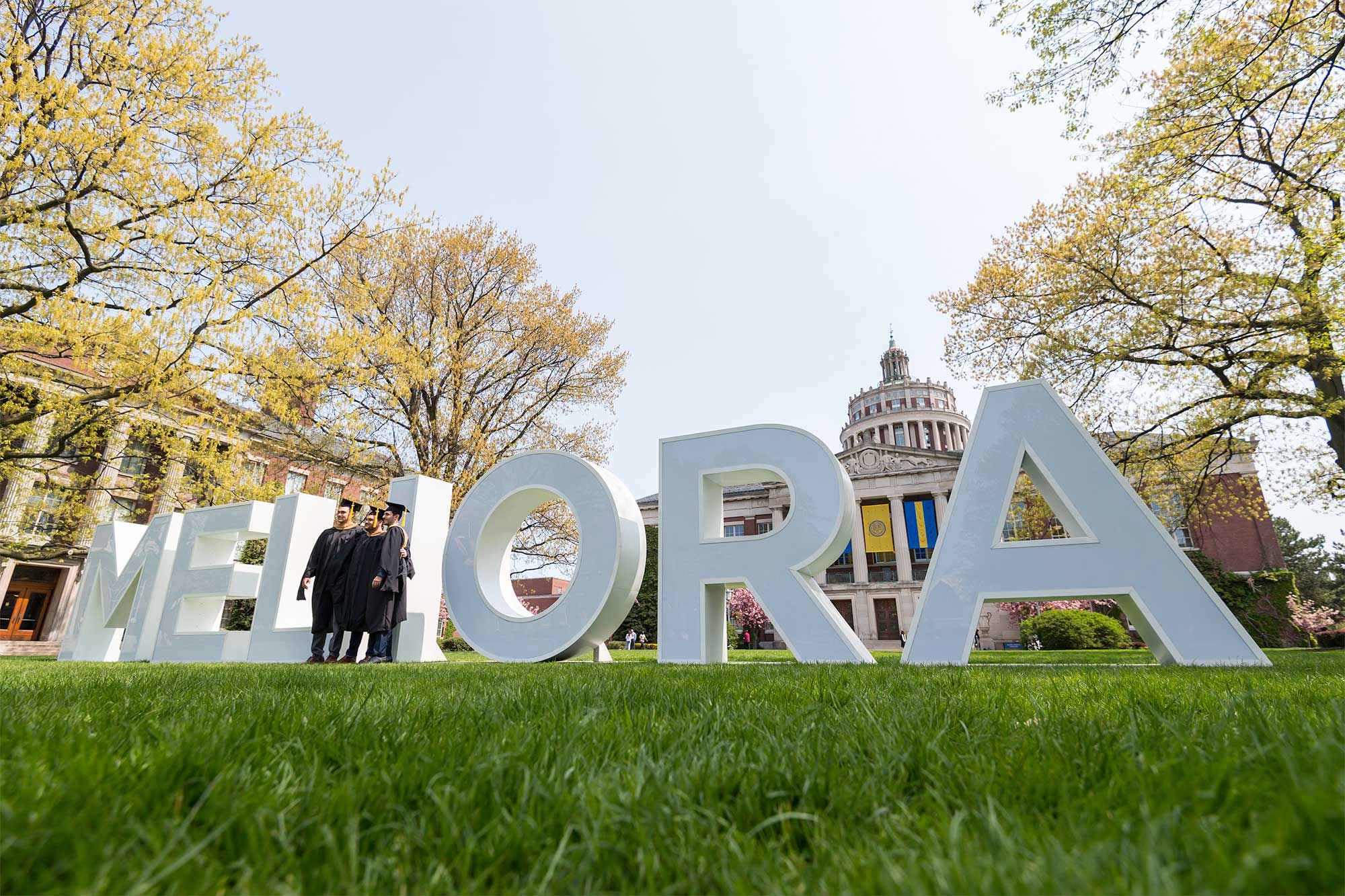 graduates in cap and gown posing in front of large white MELIORA letters in quad