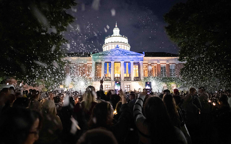 Students cheer and hold up phones to record in front of a lit-up Rush Rhees Library during Candlelight Ceremony