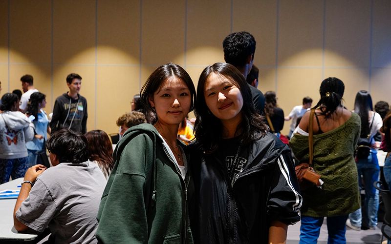 Two students smile and pose for the camera while other students mill around and chat in the background
