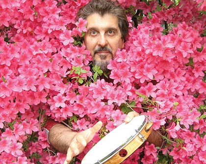 Cyro Baptista playing a tamborine in a flower hedge