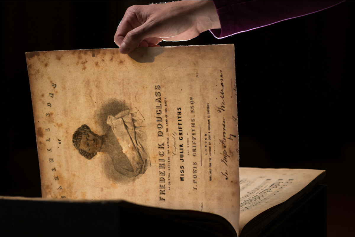 a hand holds up a sheet of music with an image of Frederick Douglass and the title FAREWELL SONG TO FREDERICK DOUGLASS 