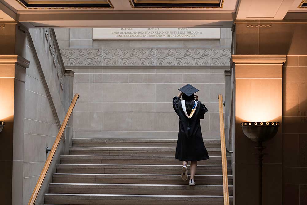 Student walks up stairs of Rush Rhees Library in cap and gown