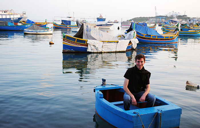 Eli Rogers smiling for the camera from a small boat with other colorful boats in the background.