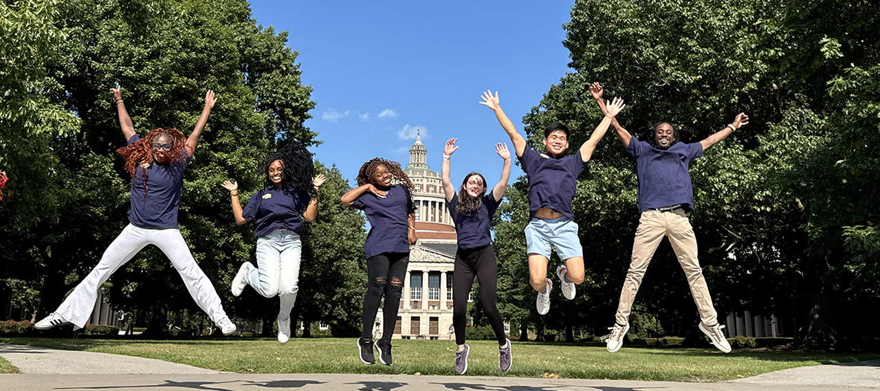 Peer advisors jumping in the air with the tower of REush Rhees and the Eastman Quad in the background.