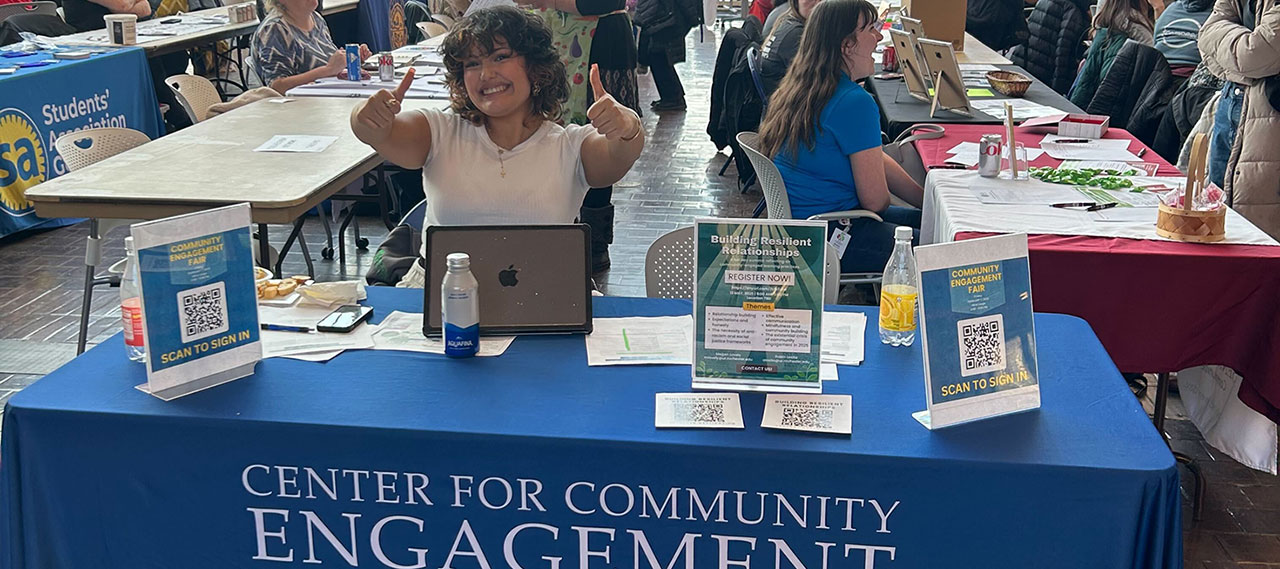 A student volunteer holding two thumbs up while sitting at a Center for Civic Engagement table during an information session.