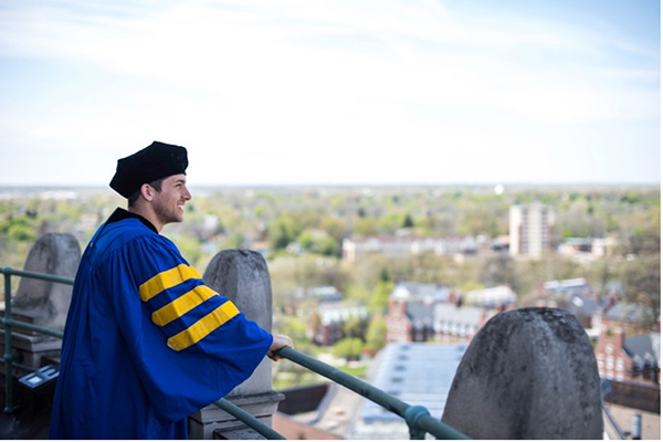 A student wearing a cap and gown is standing on the balcony at the top of Rush Rhees Library overlooking the city.