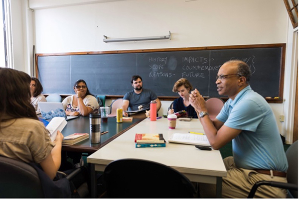 A professor and a group of students having a discussion while sitting at a table in a classroom.