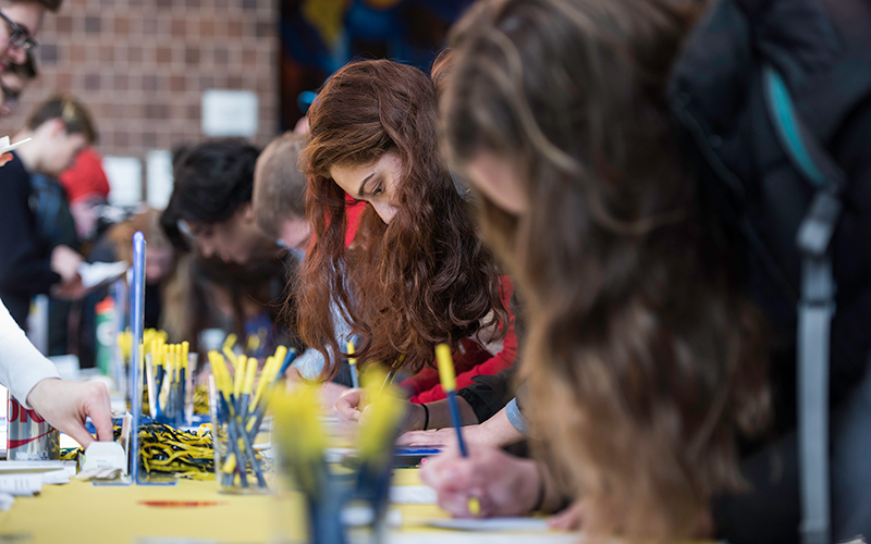 Photo of students leaning over a table and writing with blue and yellow pens