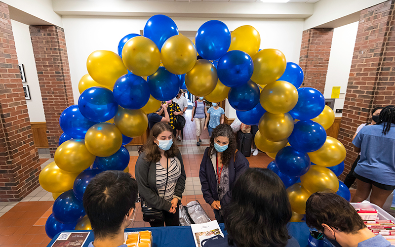 Photo of a parent and student talking to staff at a check-in table surrounded by a blue and gold balloon arch