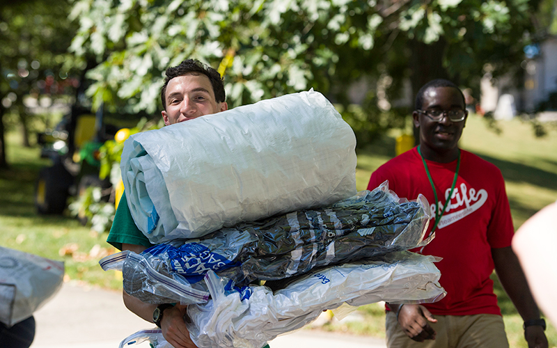 Photo of a student carrying bedding and smiling for the camera with a Residential Life student staff member in the background