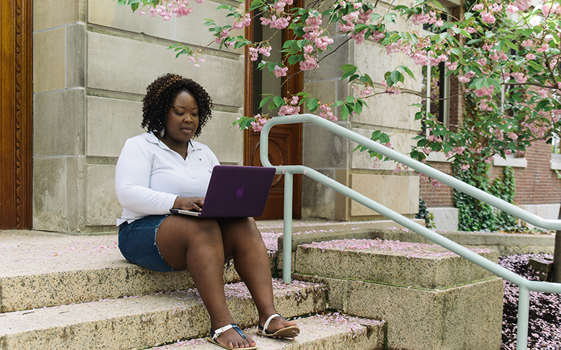 Photo of a student sitting on the steps of a building on campus looking at their laptop on their lap