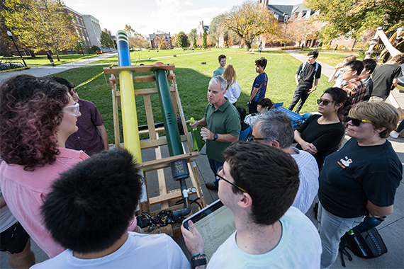 A crowd of students, faculty, and staff gather around a pressurized cannon designed to launch pumpkins.
