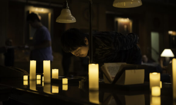 A man leans over a medieval manuscript in a dark room lit by electric candles 