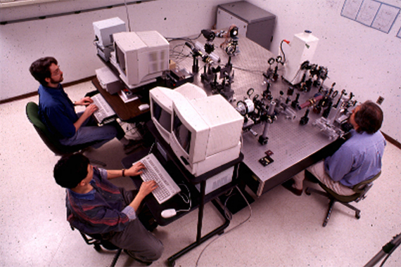 David Williams, Don Miller, and Junzhong Liang work at a table with optics equipment and computers from the 1990s.