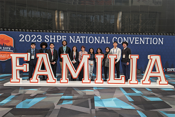 Ten Rochester students stand behind large letters that spell "FAMILIA" and in front of a banner that says "2023 SHPE NATIONAL CONVENTION"