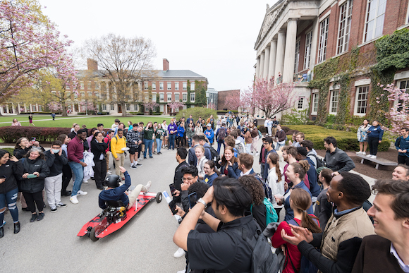 A college student drives a hand-made car powered a power drill, surrounded by a crowd of students cheering him