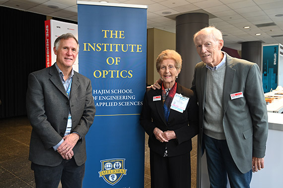 Tom Moore, Tina Kidger, and Emery Moore pose in front of a step and repeat banner that says "THE INSTITUTE OF OPTICS HAJIM SCHOOL OF ENGINEERING & APPLIED SCIENCES"