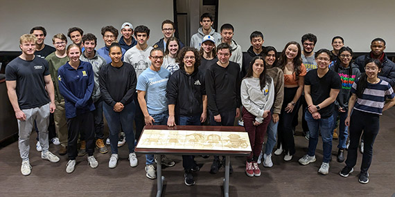 A group of nearly 30 mechanical engineering students stands in front of a jigsaw puzzle they created.