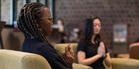 Two students meditate with their hands together in a lounge area