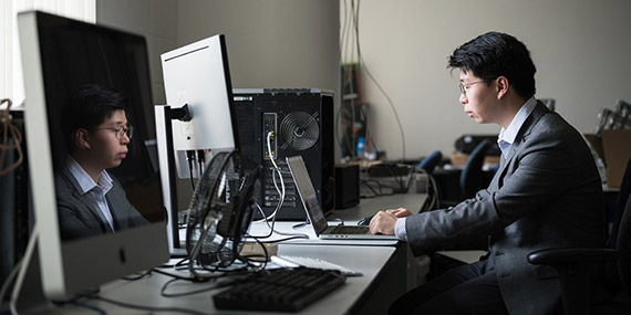 Neil Zhang works at a laptop on a desk filled with computer equipment, his face is reflected in the large monitor to his left.