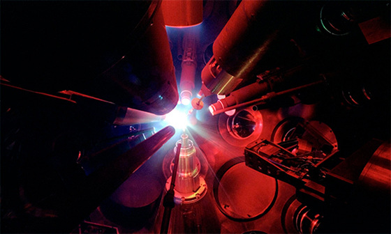 Inside the OMEGA target chamber, multiple lasers are aimed at a target amid a red cast of light.