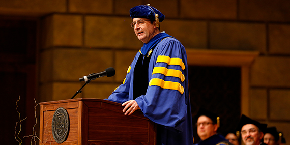 Danny Sabbah speaking at a lectern while wearing blue and yellow commencement regalia.