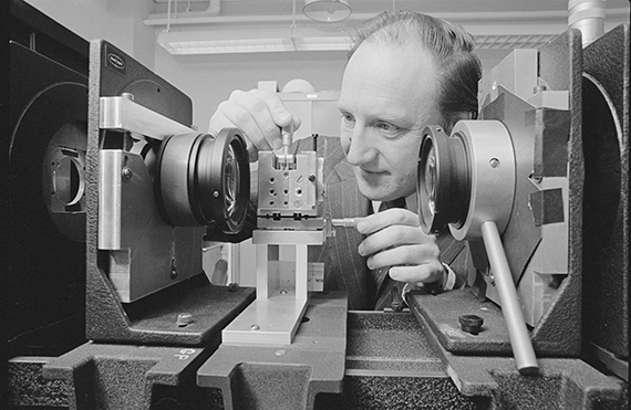 A black and white photo of Brian Thompson using optics equipment in a lab.