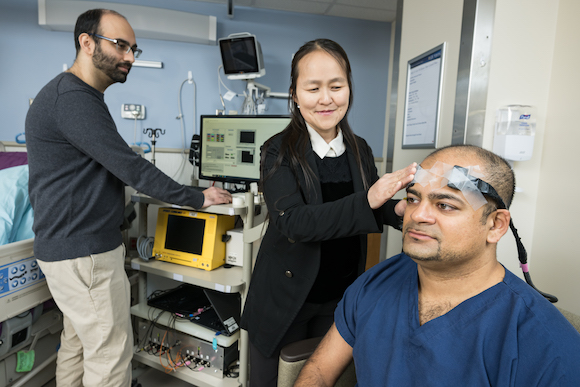 Regine Choe (center) applies diffuse correlation spectroscopy (DCS) sensors to the head of Assistant Professor Amad Khan (right) while biomedical engineering PhD student Irfan Dar (left) examines data. 