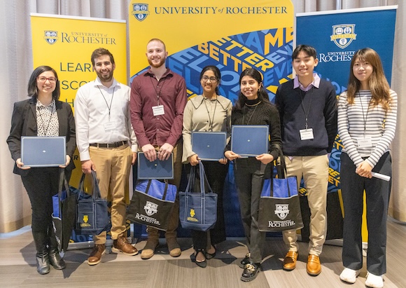 seven graduate students stand side by side in front of University of Rochester banners.