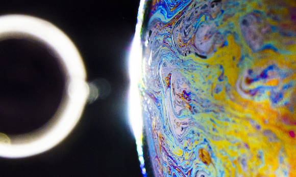 The “planet” in the artwork is a soap bubble illuminated by an extended diffuse white light. Its iridescent colors are a product of thin film interference and the striking patterns are fluid