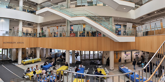 Crowds of people eat at tables and huddle around posters at Goergen Hall.