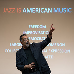 Paul BUrgett teaching History of Jazz, standing in front of a classroom screen that reads JAZZ IS AMERICAN MUSIC