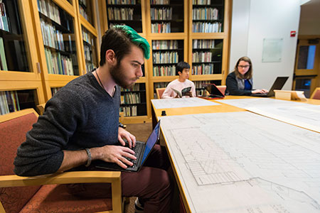 student with laptop looks at architectural drawings