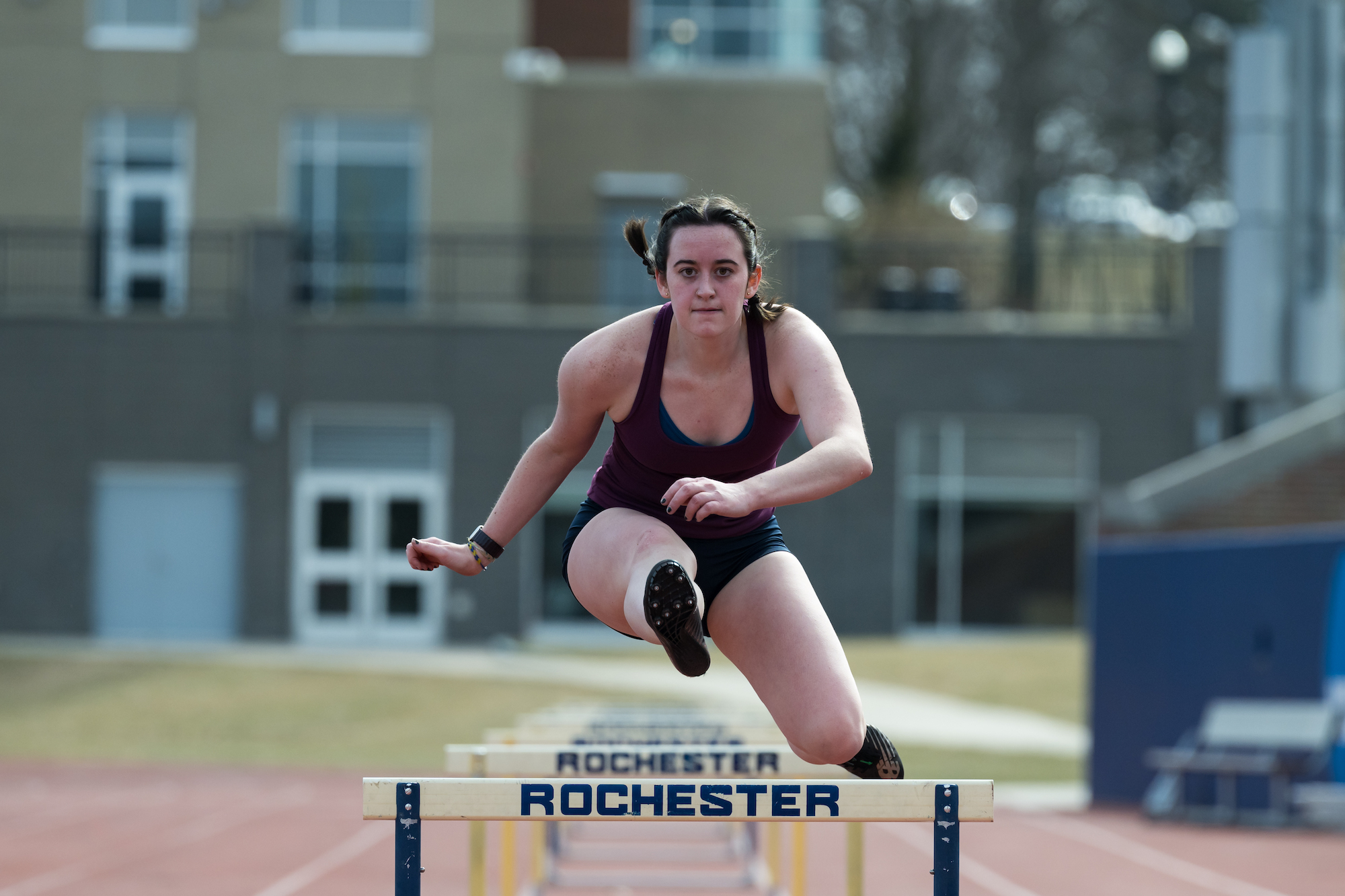 Female hurdler jumps over a Rochester-branded hurdle 