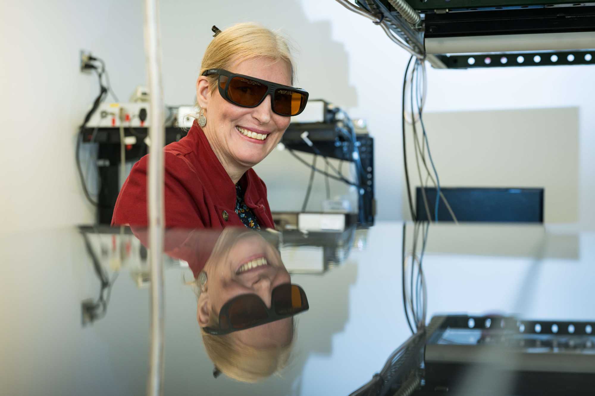 Professor and chemical engineer Astrid Muller smiling while wearing protective eyewear in her lab, with her reflection appearing in the lab equipment below her. 