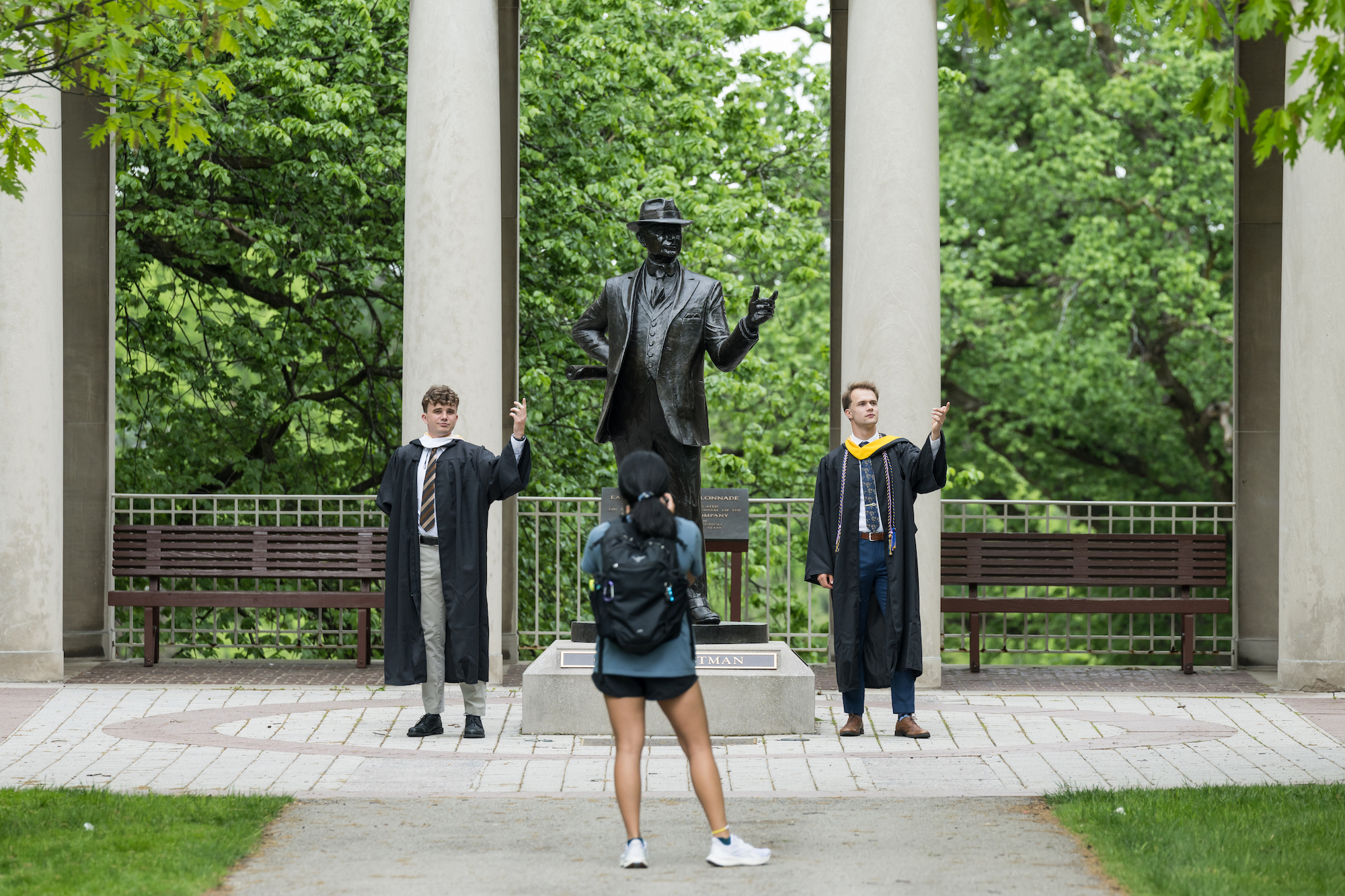 Two students in graduation regalia pose with a statue of George Eastman. Another student takes their picture