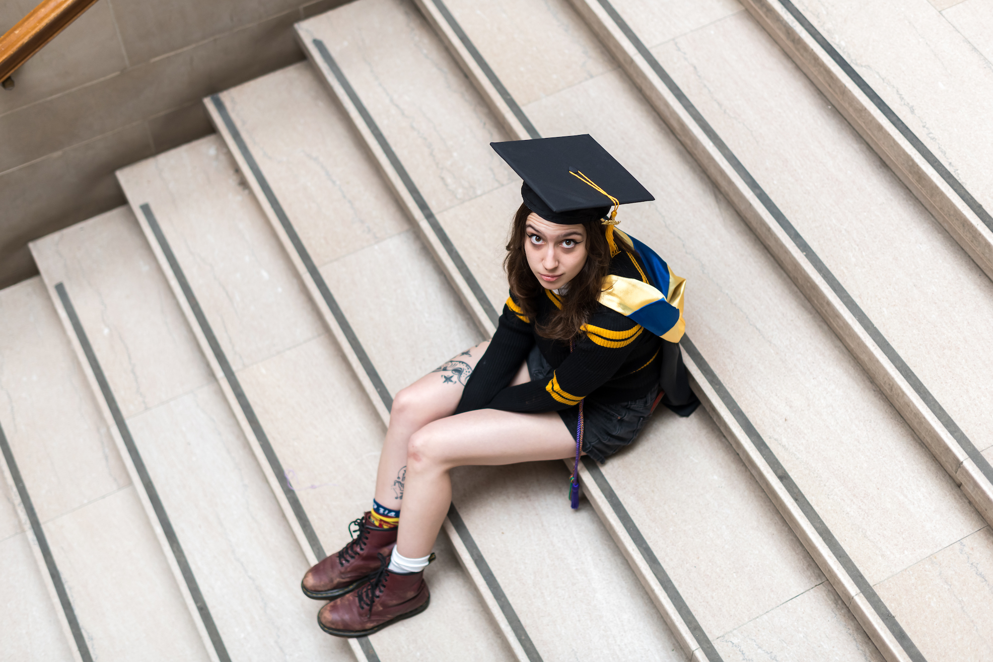 A student in graduation regalia sits on steps looking up at the camera