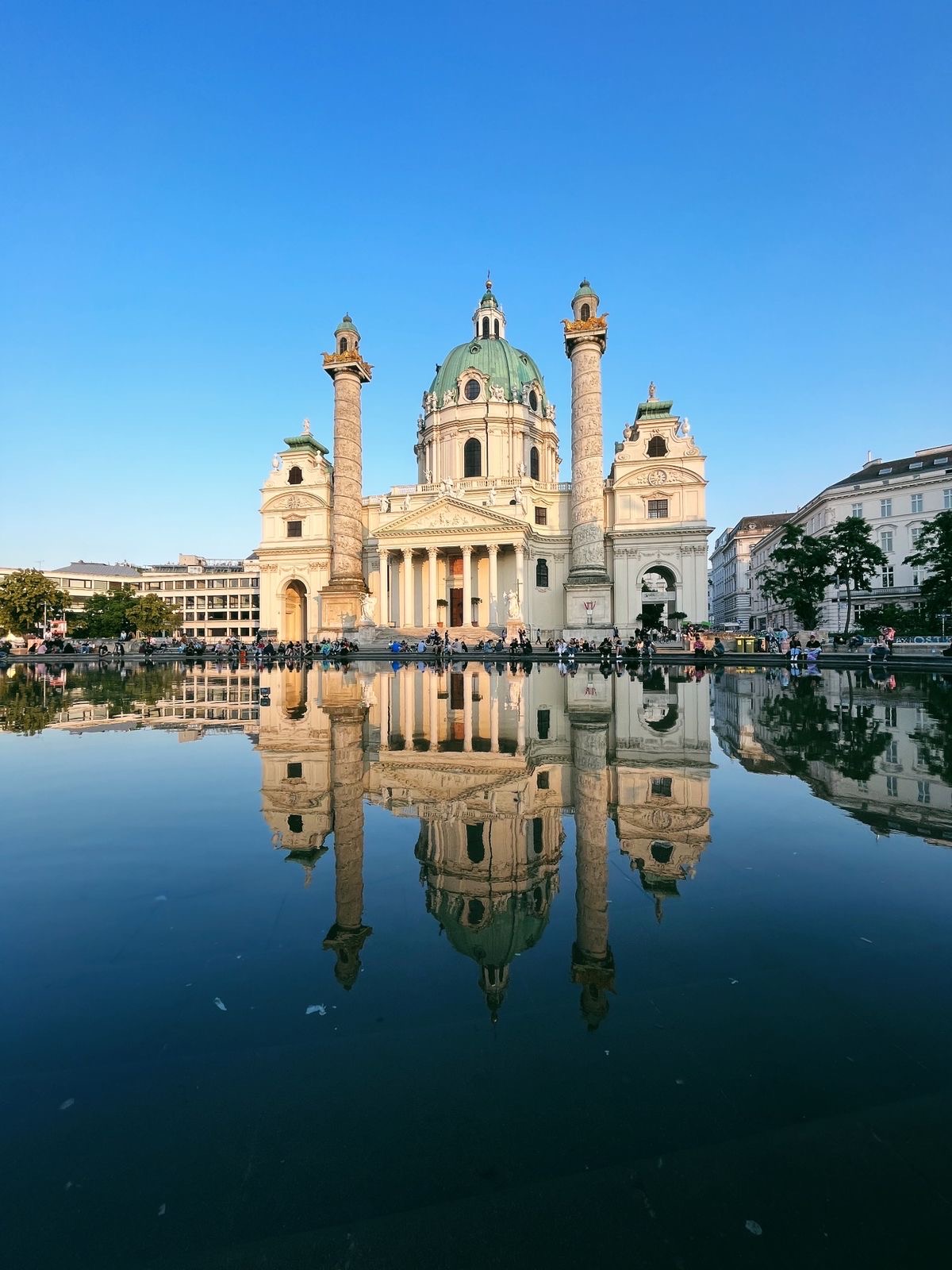 A grand baroque-style white cathedral with a large green dome and twin columns stands against a clear blue sky. The building is beautifully reflected in a calm, glassy pool of water in the foreground, with people gathered around its base.