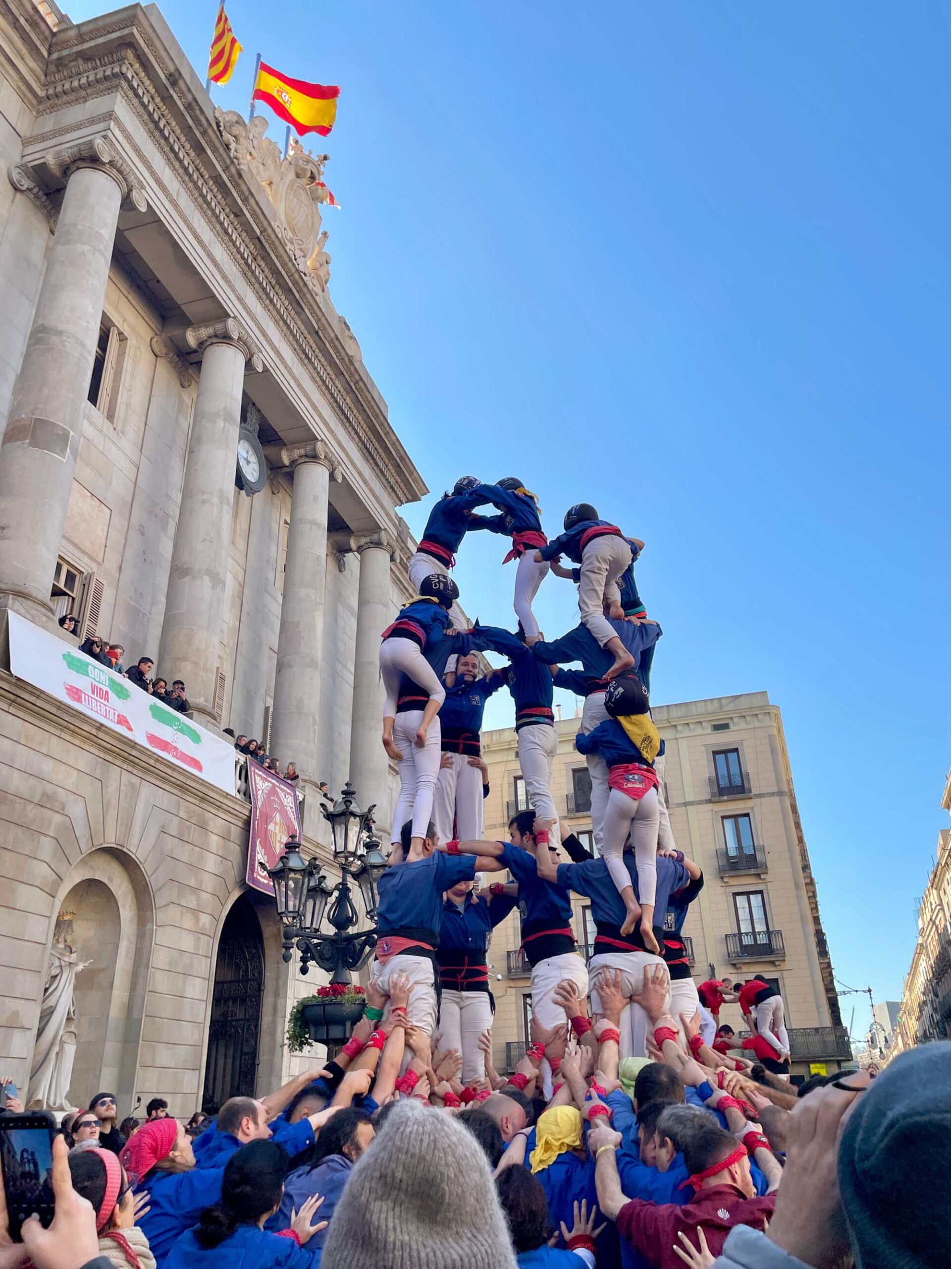 A human tower is formed by a group of people, standing in front of a historic building with columns. Flags are seen atop the building. The base of the tower is wide, gradually narrowing as it reaches the top, with several tiers of participants. Onlookers are gathered around.