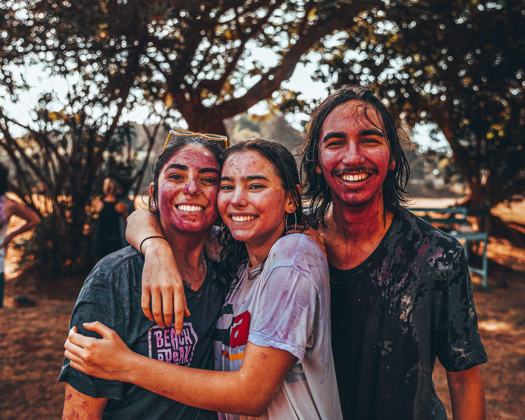 Three young people covered in colorful powder stand outdoors, smiling and hugging, with trees in the background.