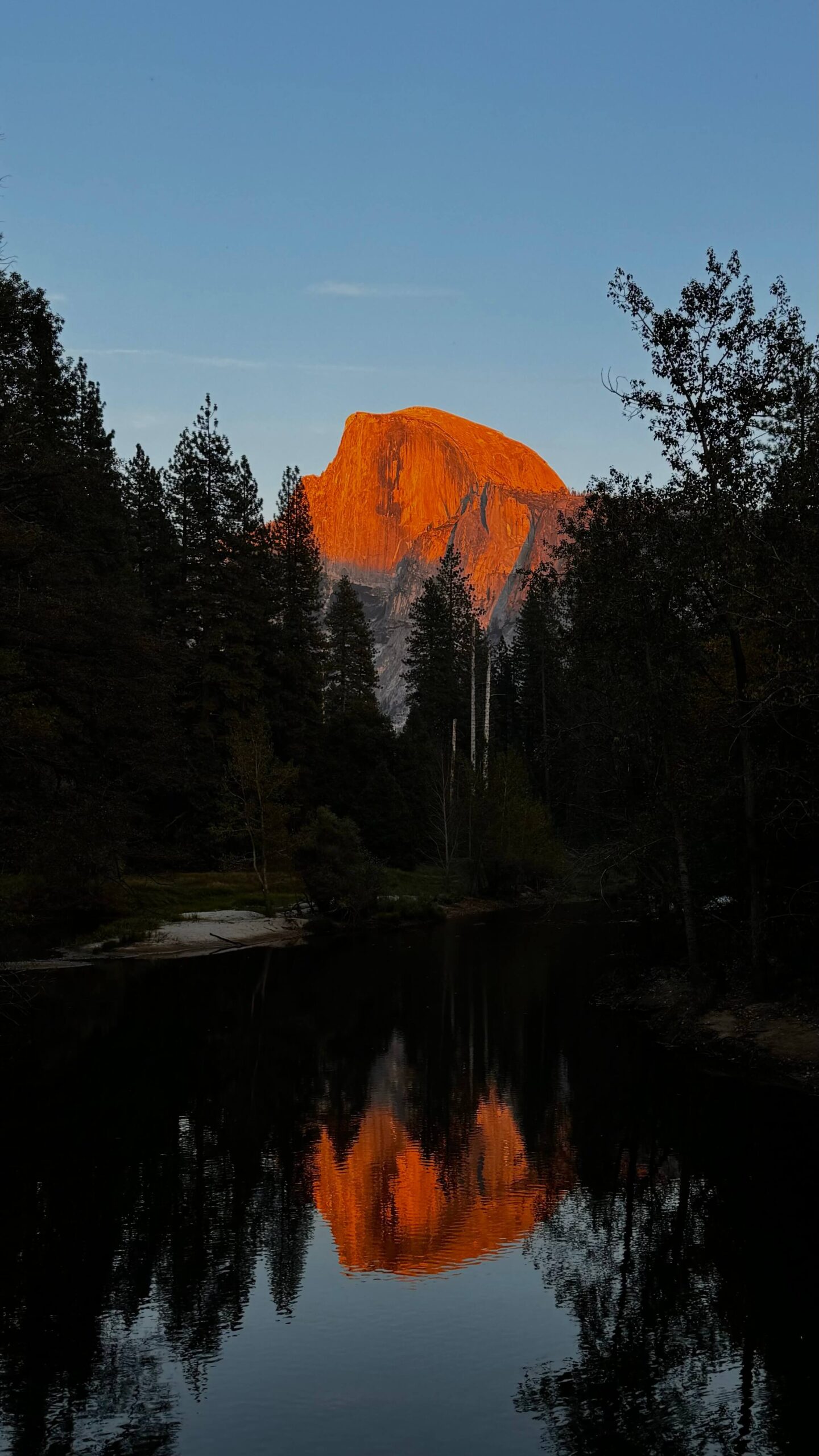 A striking orange sunset illuminates the peak of Half Dome in Yosemite National Park, framed by tall trees. The clear, still water below mirrors the vibrant rock formation against a deepening blue sky.