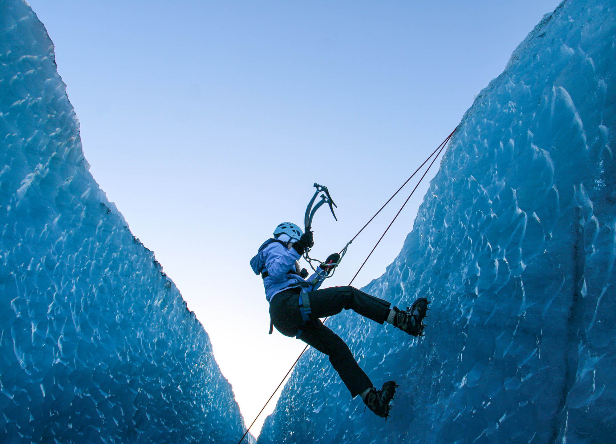 A person wearing climbing gear, including a helmet and crampons, is ice climbing between two tall ice walls. They are using ice axes and are secured by ropes. The sky in the background is clear and bright.