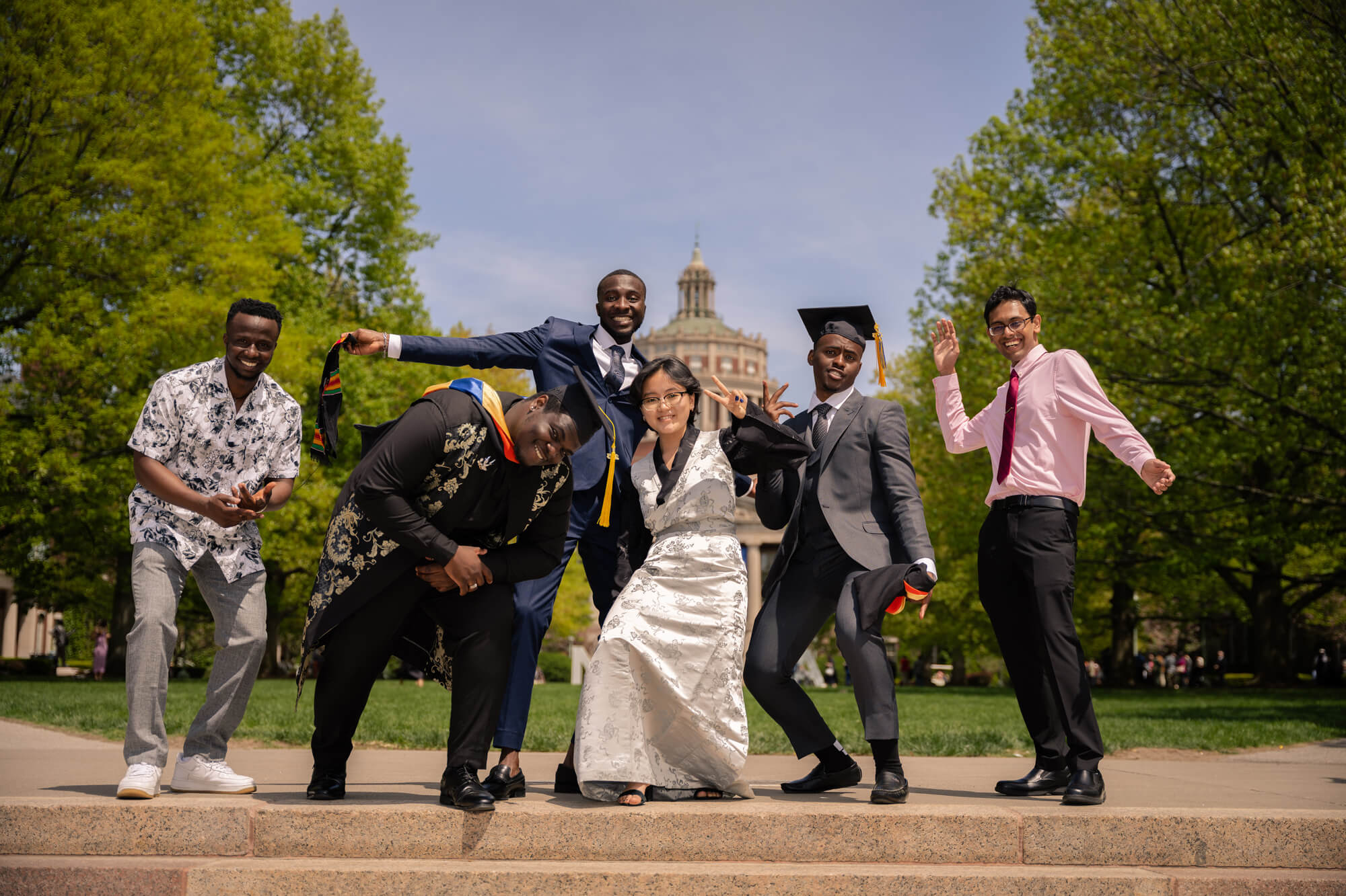 A group of six diverse graduates, dressed in caps, gowns, and formal wear, joyfully pose for a celebratory photo on a sunny day in front of a historic building surrounded by lush green trees. They are smiling and striking playful poses, exuding excitement and pride.