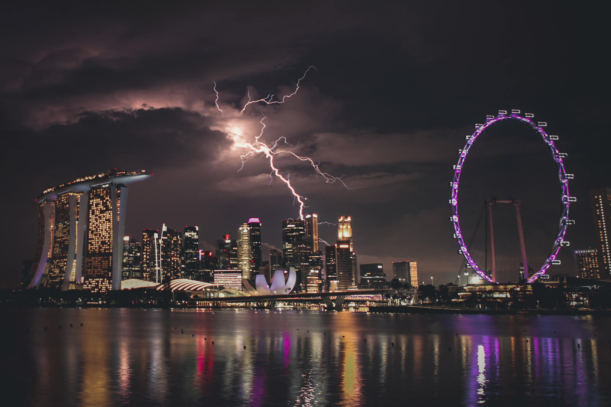 A vibrant nighttime cityscape of Singapore with lightning striking the sky. The illuminated skyline features the Marina Bay Sands hotel, the lotus-shaped ArtScience Museum, and the glowing Singapore Flyer Ferris wheel, all reflecting on the tranquil water.
