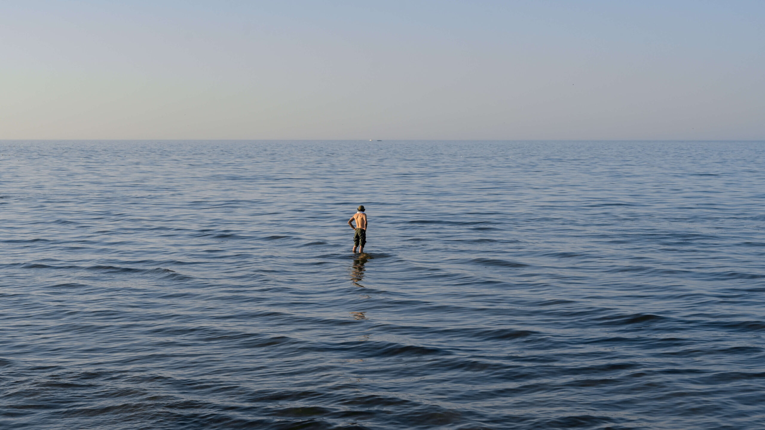 A person stands in the expansive, calm sea under a clear sky, with water stretching to the horizon. Their reflection is visible on the water's surface, creating a tranquil and serene scene.
