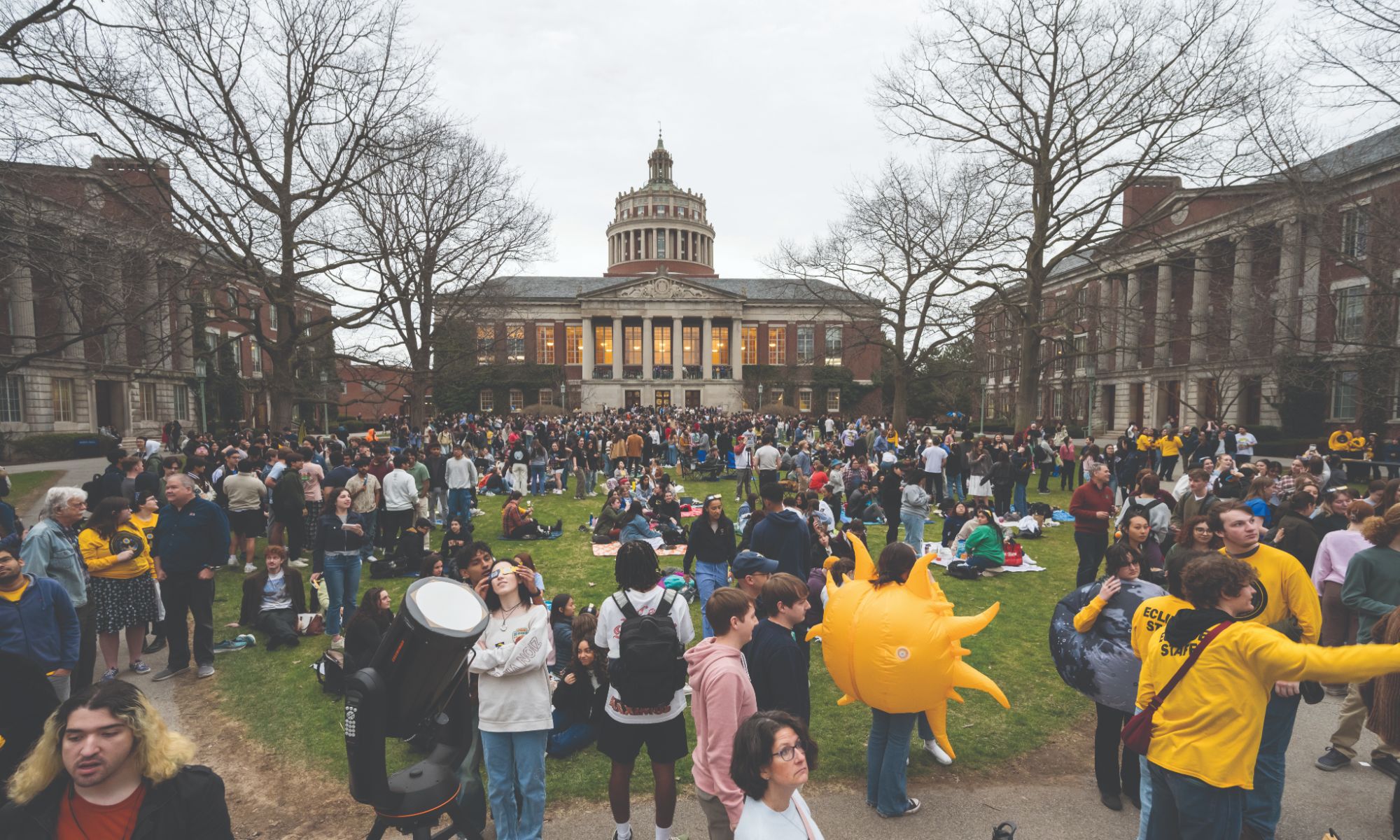 A bustling crowd on a university campus gathers on a green lawn with a large domed building in the background. Many attendees wear yellow, and some carry inflatable suns. There is also a large telescope in the foreground. The scene is lively and energetic.