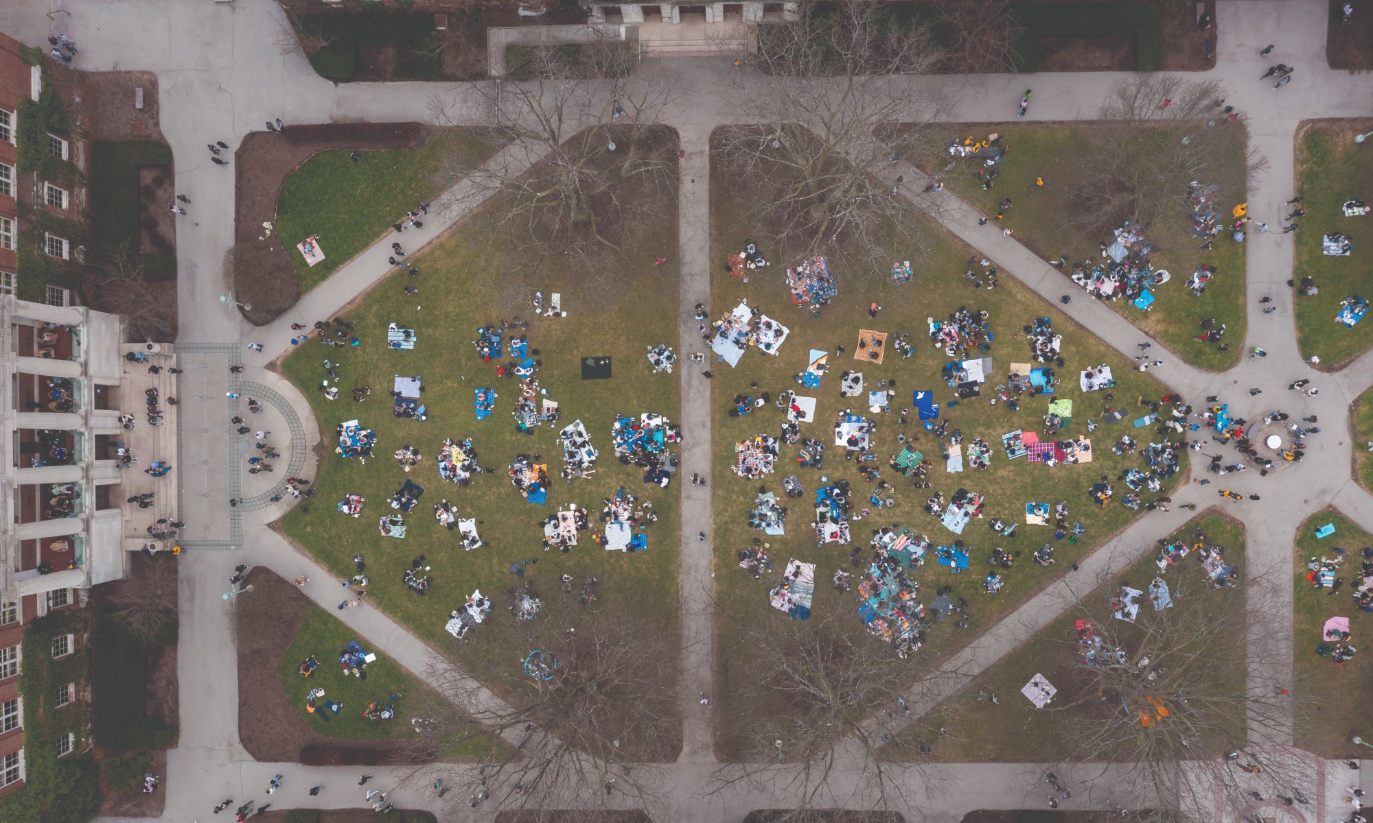 Aerial view of a large park with people gathered on blankets in groups across two main grassy areas divided by pedestrian pathways. The park is surrounded by trees and buildings, with intersecting paths creating symmetrical patterns.