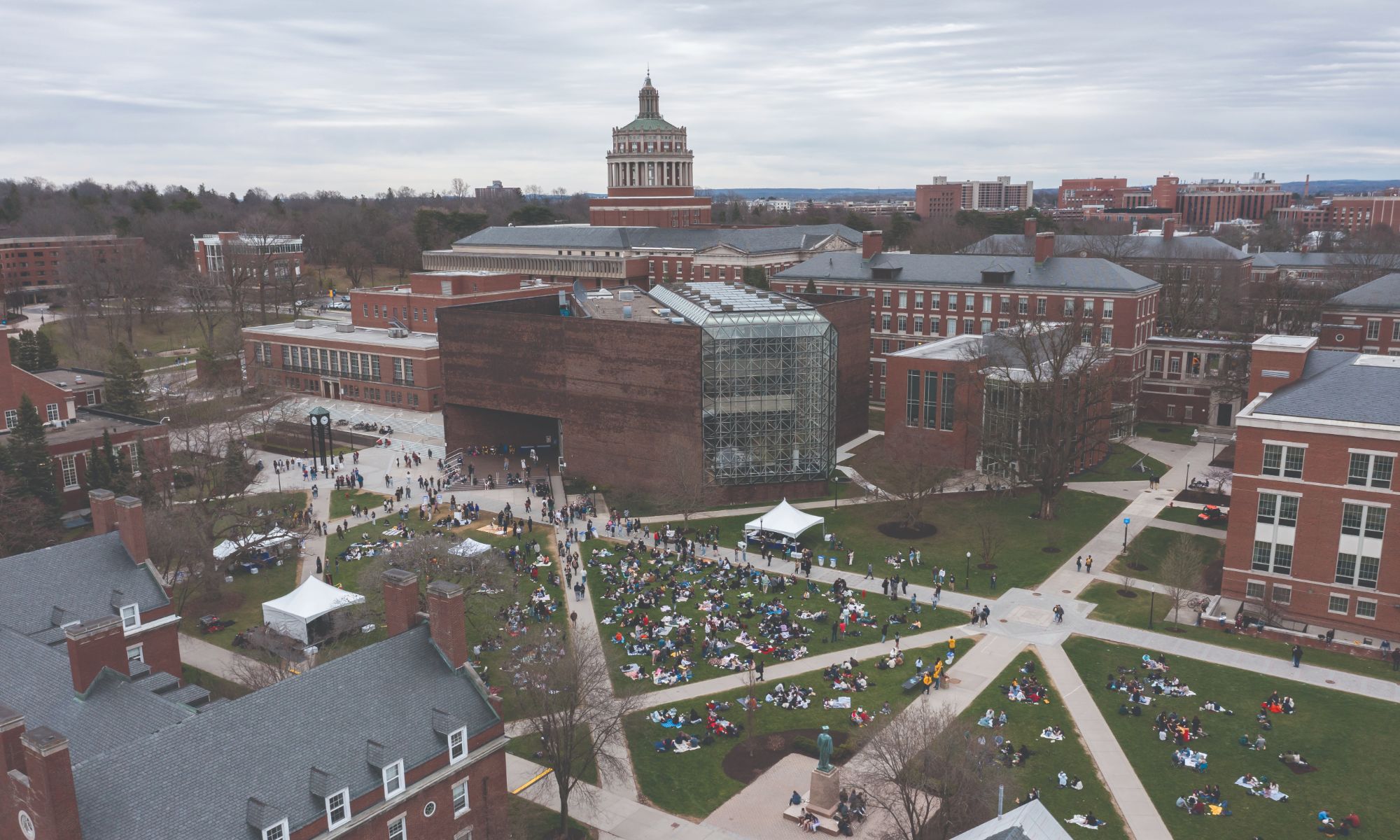Aerial view of a college campus with students gathered on lawns. The campus features several brick buildings, including one with a prominent dome. Tents and groups of people are visible, suggesting an event. The sky is overcast.