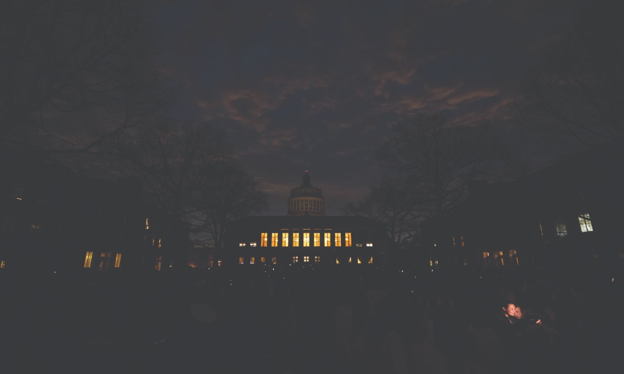 University of Rochester library is seen at dusk with its windows brightly illuminated. The sky is dark with scattered clouds, and the silhouettes of trees and people are visible in the foreground.