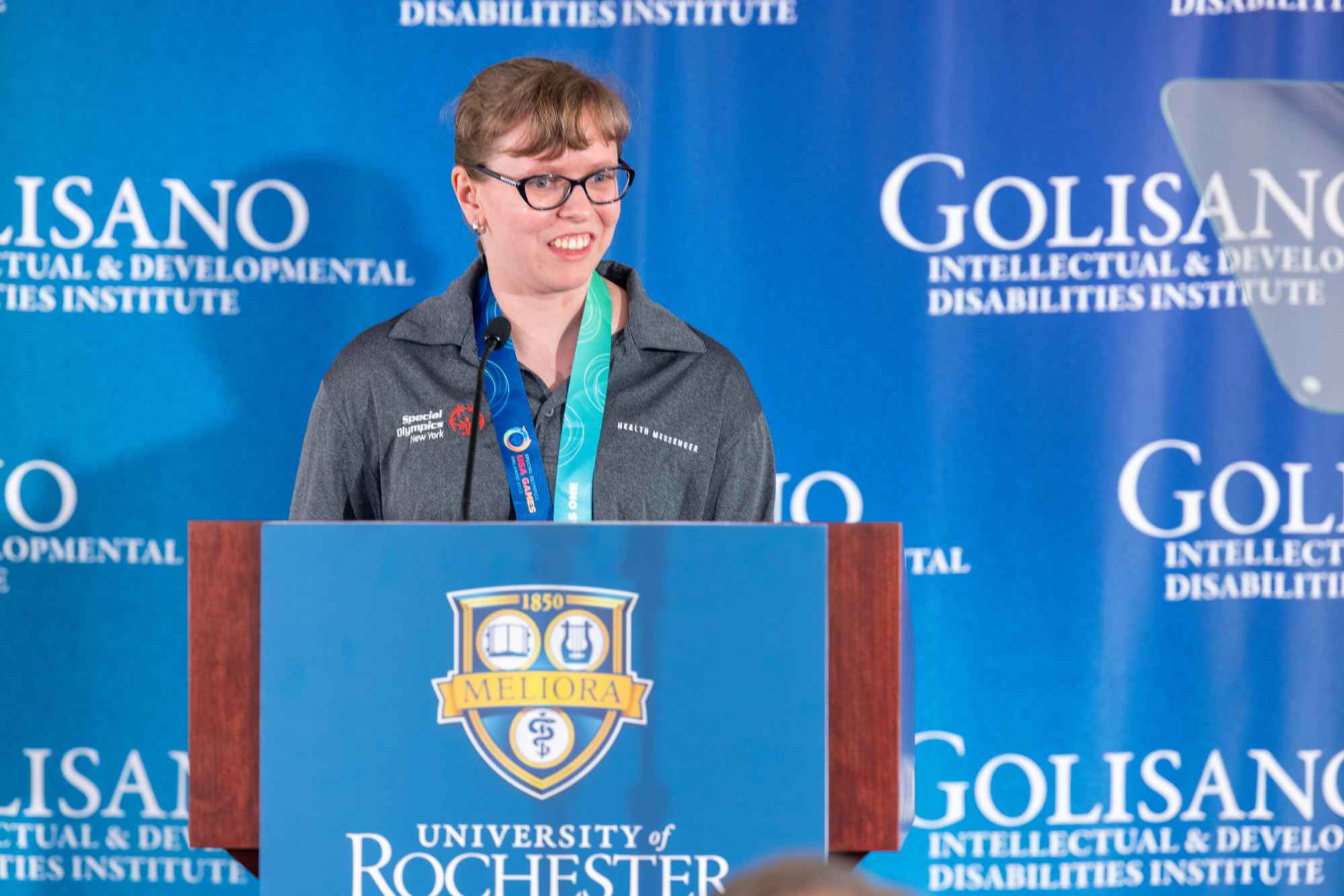 A female Special Olympics gold medalist and ambassador stands at a University of Rochester podium in front of a blue backdrop for the Golisano Intellectual and Developmental Disabilities Institute. 